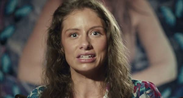 A person with long hair is speaking directly to the camera with an expressive facial expression, wearing a patterned top