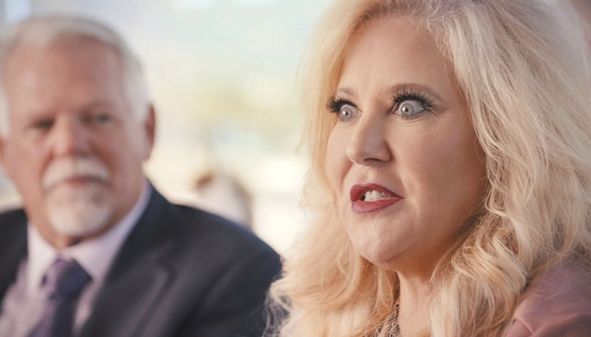 Two people in a conversation, a woman speaking in the foreground. Both are dressed in business attire