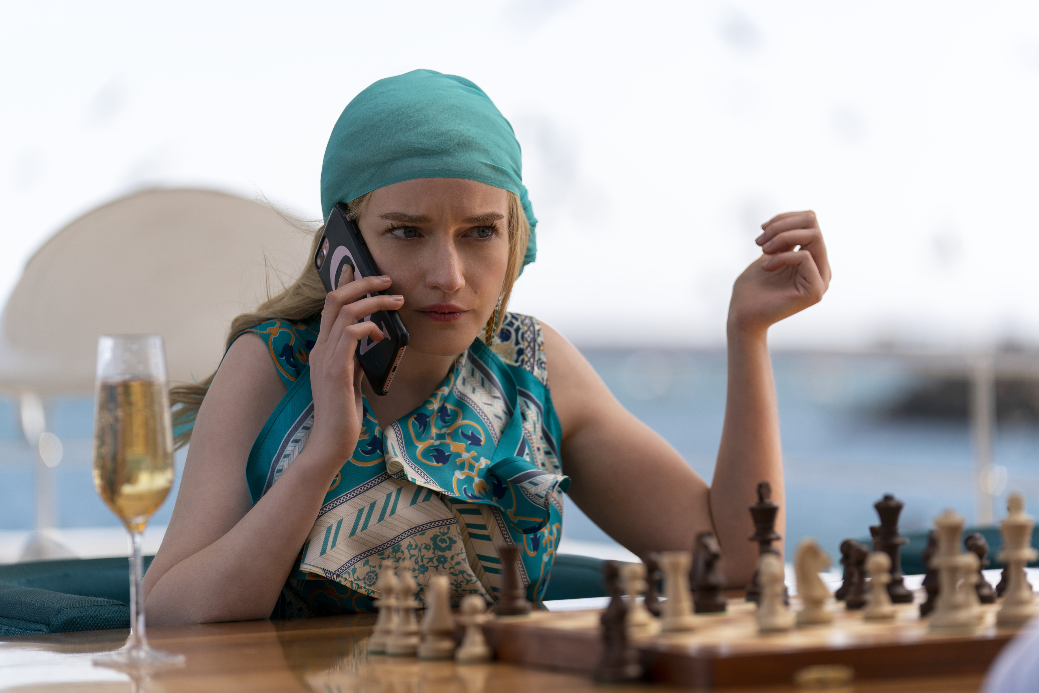 Julia Garner as Anna Delvey talking on a phone while sitting outside in front of a chessboard with a glass of champagne on the table next to her