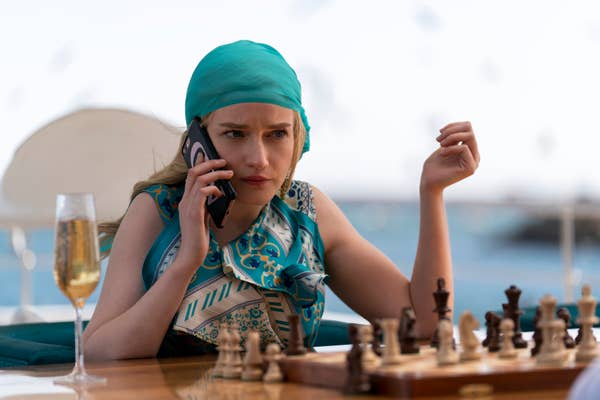 Julia Garner as Anna Delvey talking on a phone while sitting outside in front of a chessboard with a glass of champagne on the table next to her