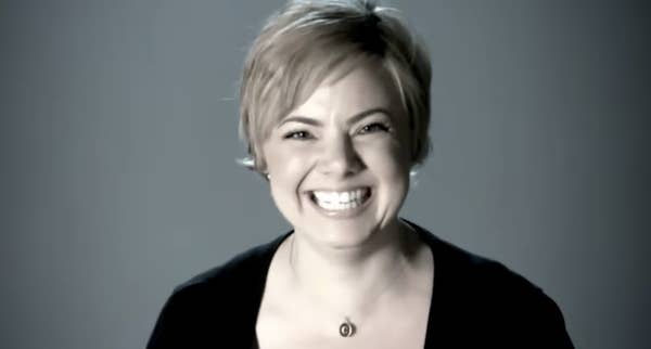 Person with short hair smiling, wearing a dark shirt, against a plain background
