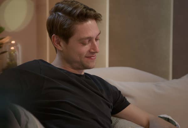 Mason is sitting on a couch, smiling softly, with a relaxed and content expression
