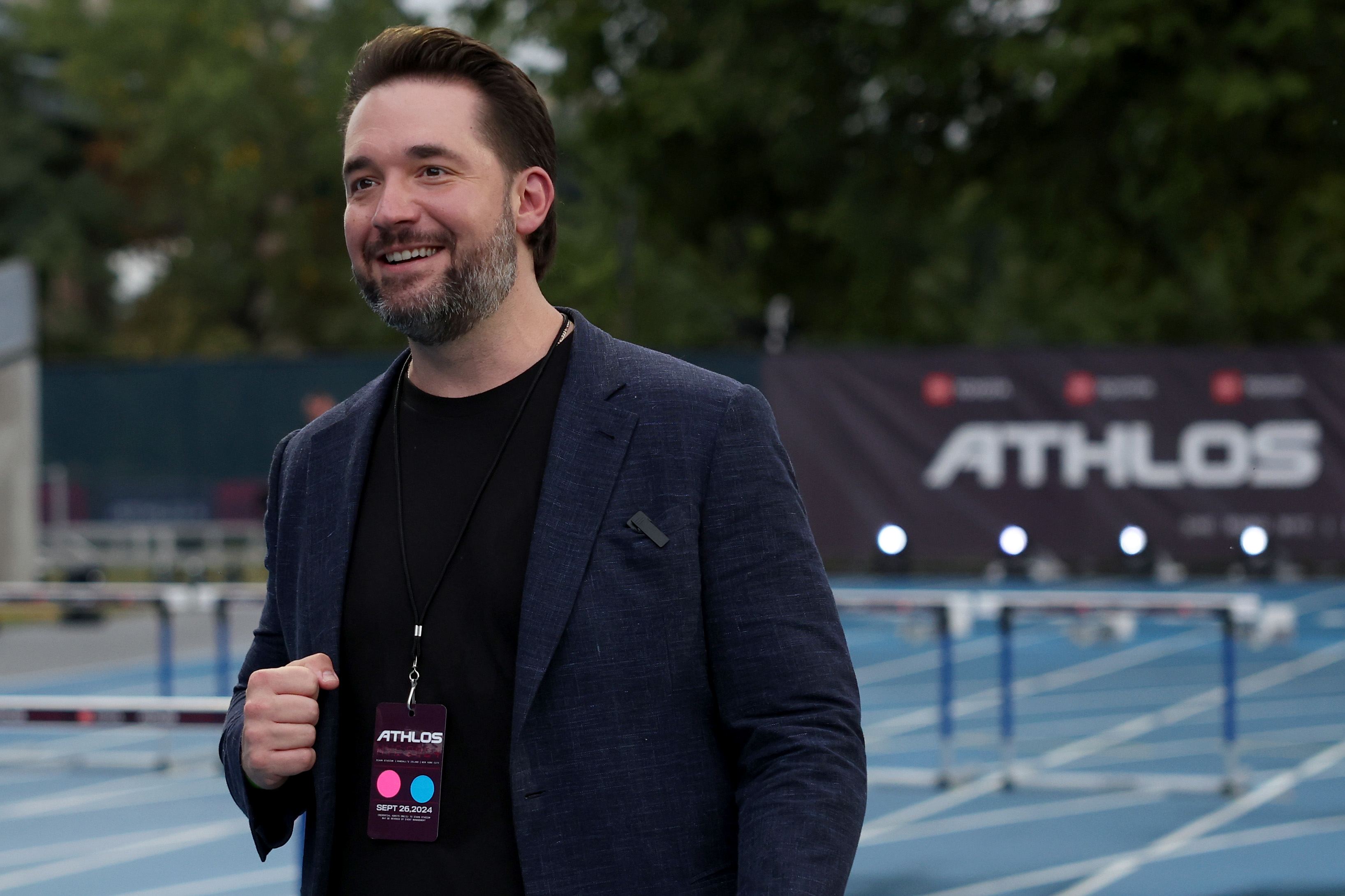 Alexis Ohanian at an outdoor event wearing a dark blazer, smiling, with a lanyard displaying a name. Background includes a track and an "ATHLOS" banner