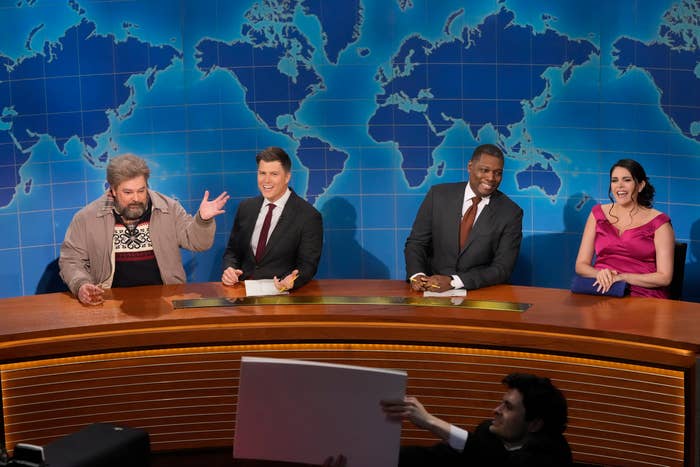 Four comedians sit at a news desk on a TV show set with a world map backdrop, engaging in a lively discussion. One person holds up a cue cards