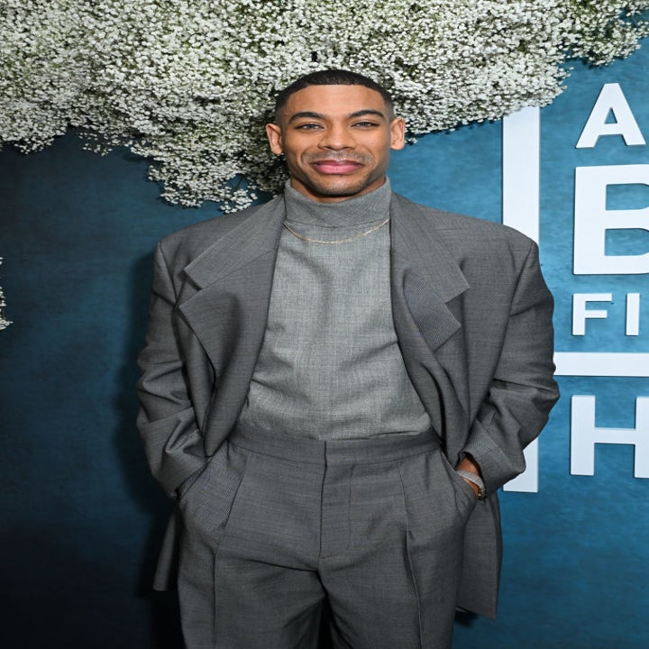 Aaron in a stylish suit poses against a backdrop of flowers and a wall with partial text