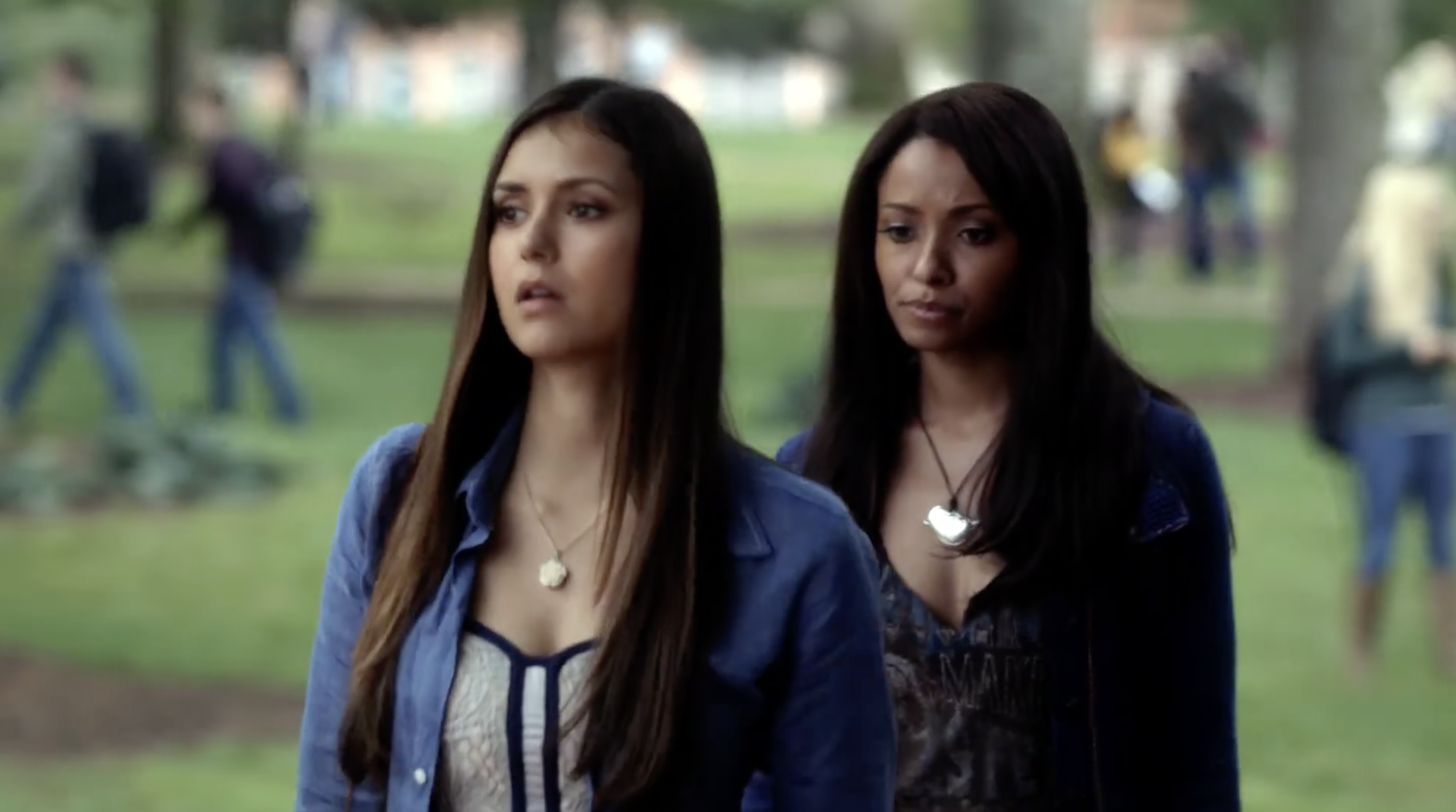 Two women walking outdoors in a park, both wearing casual outfits with open jackets. They appear serious and focused, looking ahead