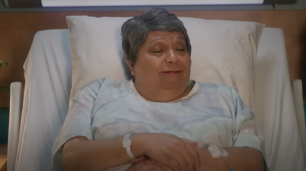 Person lying in a hospital bed, wearing a hospital gown, with wristbands visible. Appears relaxed, eyes gently closed, and a slight smile