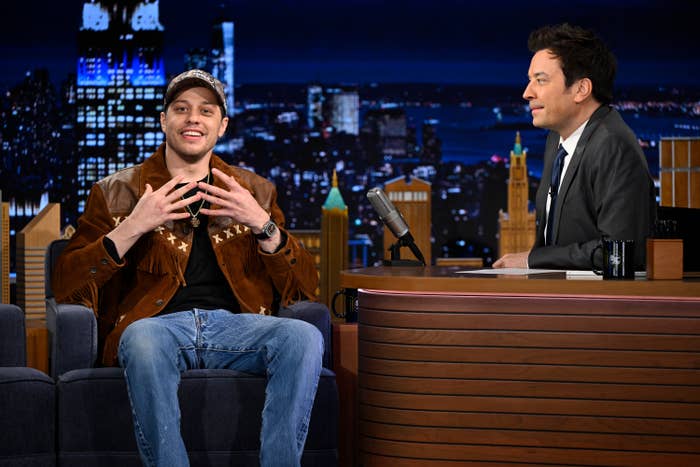 Two men sit and talk on a late-night talk show set. One wears a casual jacket and jeans, the host wears a suit