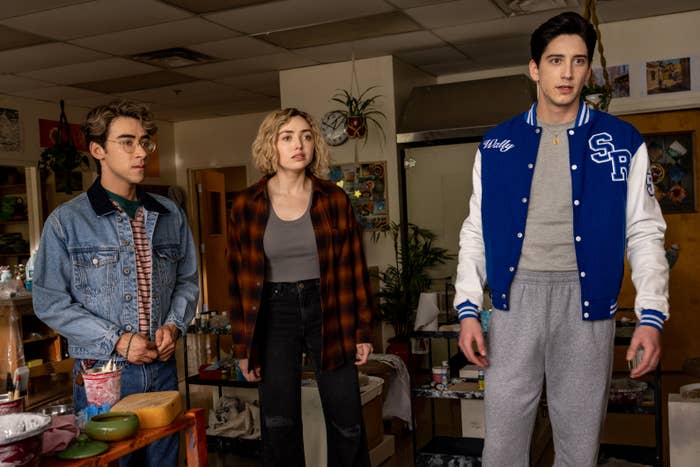 Three young adults stand indoors; one wears a denim jacket, one a plaid shirt, and the third a blue varsity jacket. They appear surprised in a scene from &quot;School Spirits&quot;