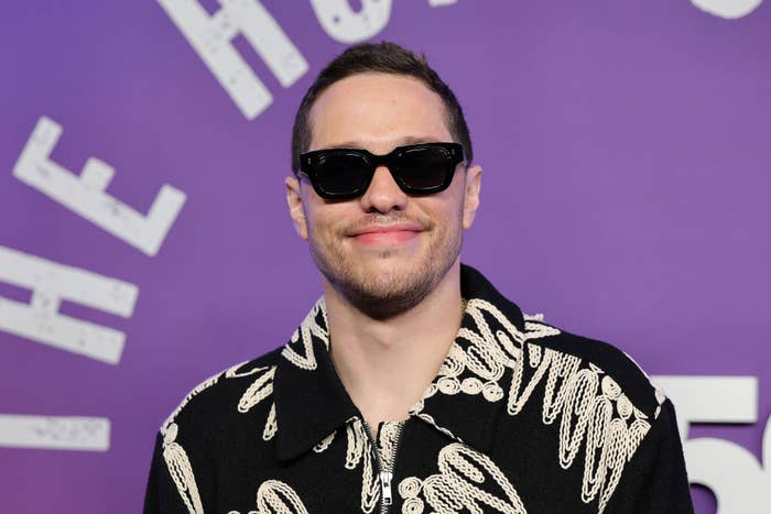 Person wearing sunglasses and a patterned jacket at an event with a purple backdrop