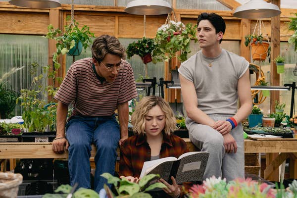 Three young adults in casual attire are in a greenhouse, with one reading a notebook, surrounded by plants