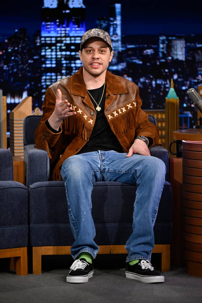 Person wearing a fringed jacket, baseball cap, jeans, and sneakers, sitting on a talk show set, gesturing while speaking