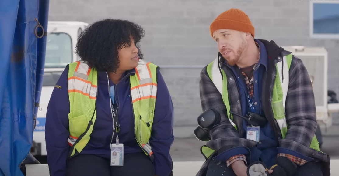 Two people talking, one wearing a knit hat and work vest, looking engaged in conversation