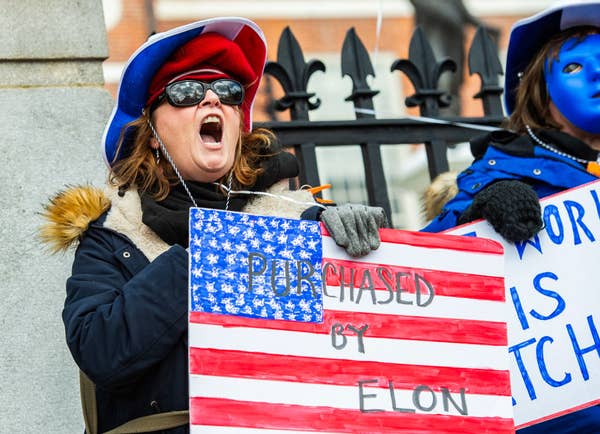A protester shouts while holding a sign with U.S. flag imagery that reads "Purchased by Elon" at a public demonstration