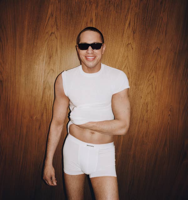 Person in fitted white t-shirt and shorts, wearing sunglasses, smiling against a wooden wall