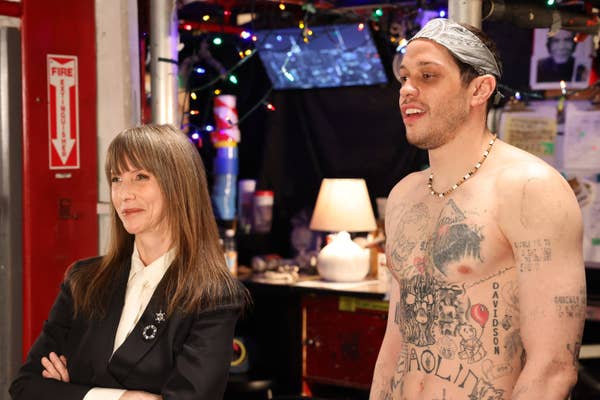 Two people backstage, one wearing a suit with a star brooch, the other shirtless with tattoos and a necklace, surrounded by stage equipment