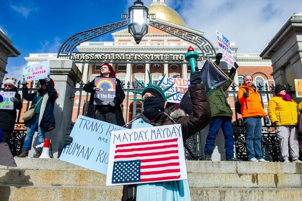 Protesters outside a building hold various signs advocating for trans rights and against wealth disparity. A person is dressed as the Statue of Liberty