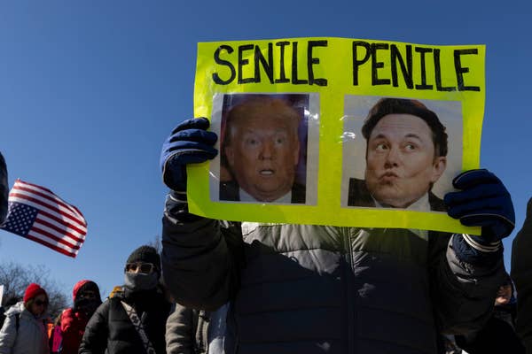 Person holding a sign with photos labeled "Senile" and "Penile" at a protest. Crowd and a U.S. flag in the background