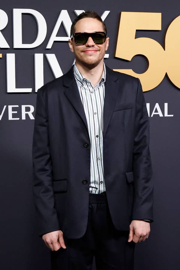 Person in a dark suit and striped shirt on a red carpet, wearing sunglasses, standing in front of a &quot;Saturday Night Live&quot; backdrop