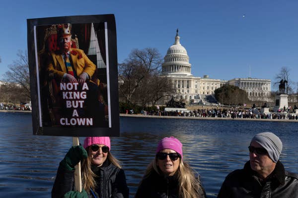 Three people hold a protest sign showing a clown king near the U.S. Capitol, with text "Not a king but a clown."