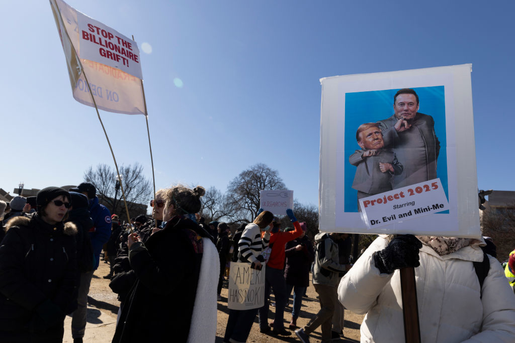 People at a protest holding signs, one depicting caricatures of Elon Musk and another figure as Dr. Evil and Mini-Me, labeled "Project 2025."