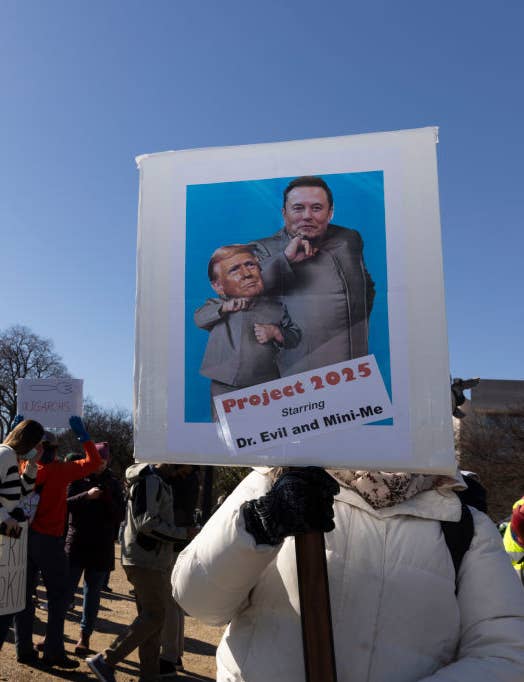 People at a protest holding signs, one depicting caricatures of Elon Musk and another figure as Dr. Evil and Mini-Me, labeled "Project 2025."
