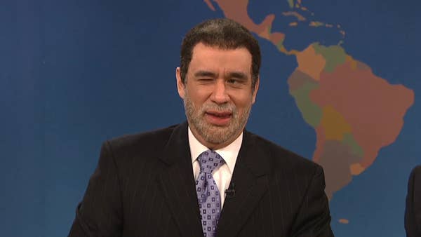 Person in suit and patterned tie winking at the camera against a map backdrop, likely part of a TV or comedy skit setting