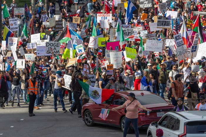 A large crowd of protesters marches with various signs, some related to immigration and politics, alongside a red car on a city street