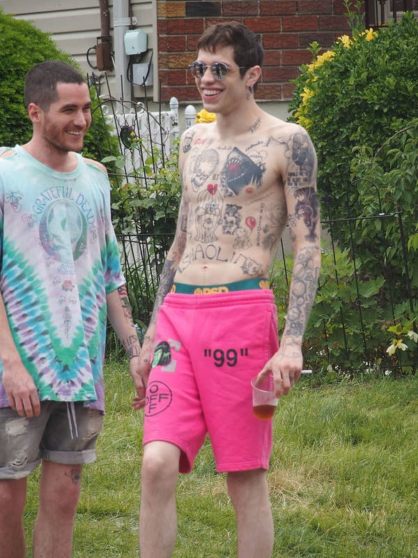 Two people outdoors; one man, tattooed and shirtless, wears bright pink shorts and sunglasses, holding a drink, smiling