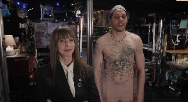 Two people stand backstage. One is smiling in a suit, the other is shirtless with tattoos, wearing a beanie