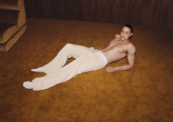 A shirtless man poses on a carpeted floor, wearing white pants and socks