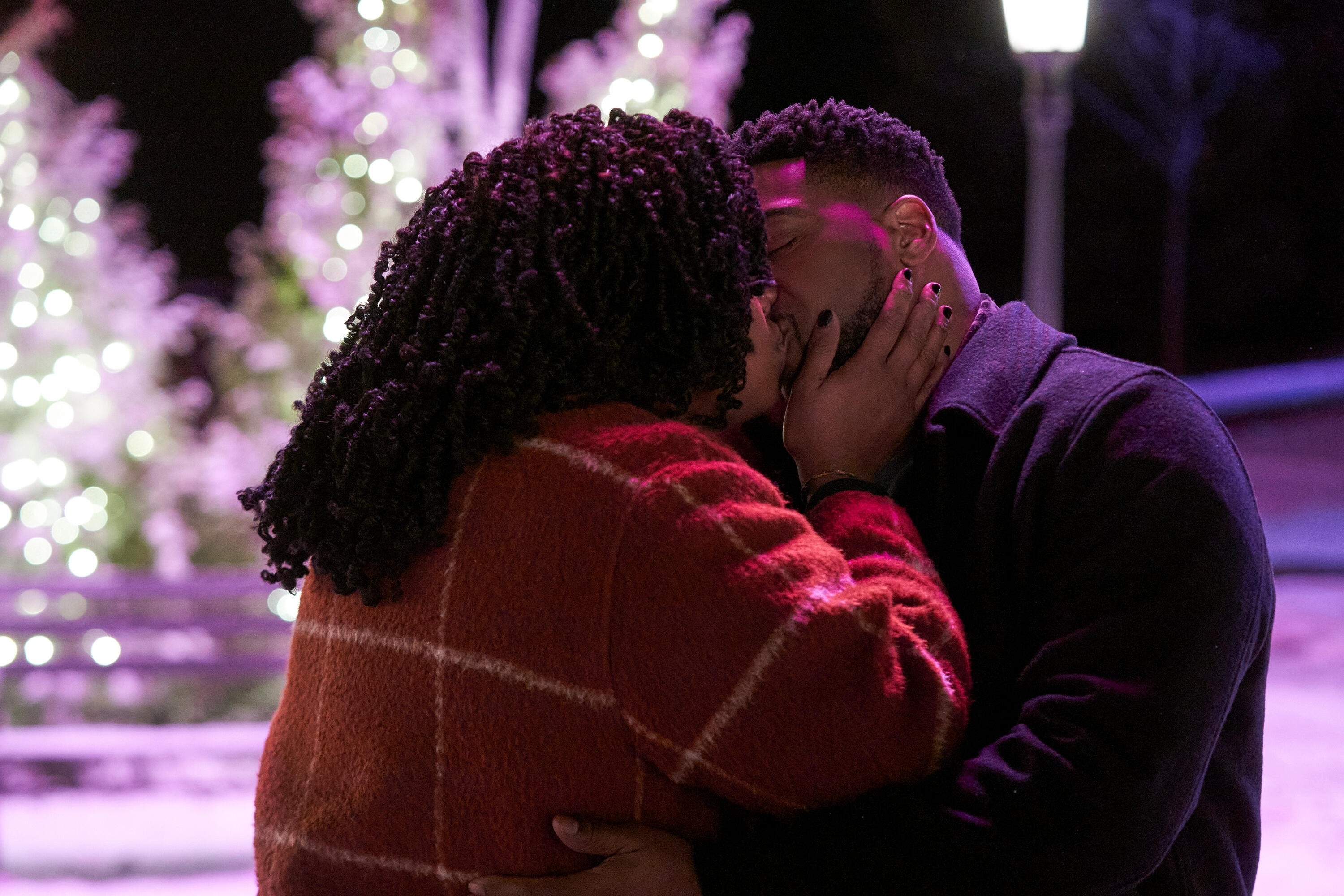 A couple shares a romantic kiss outdoors at night, surrounded by snow and illuminated trees, creating a cozy winter scene