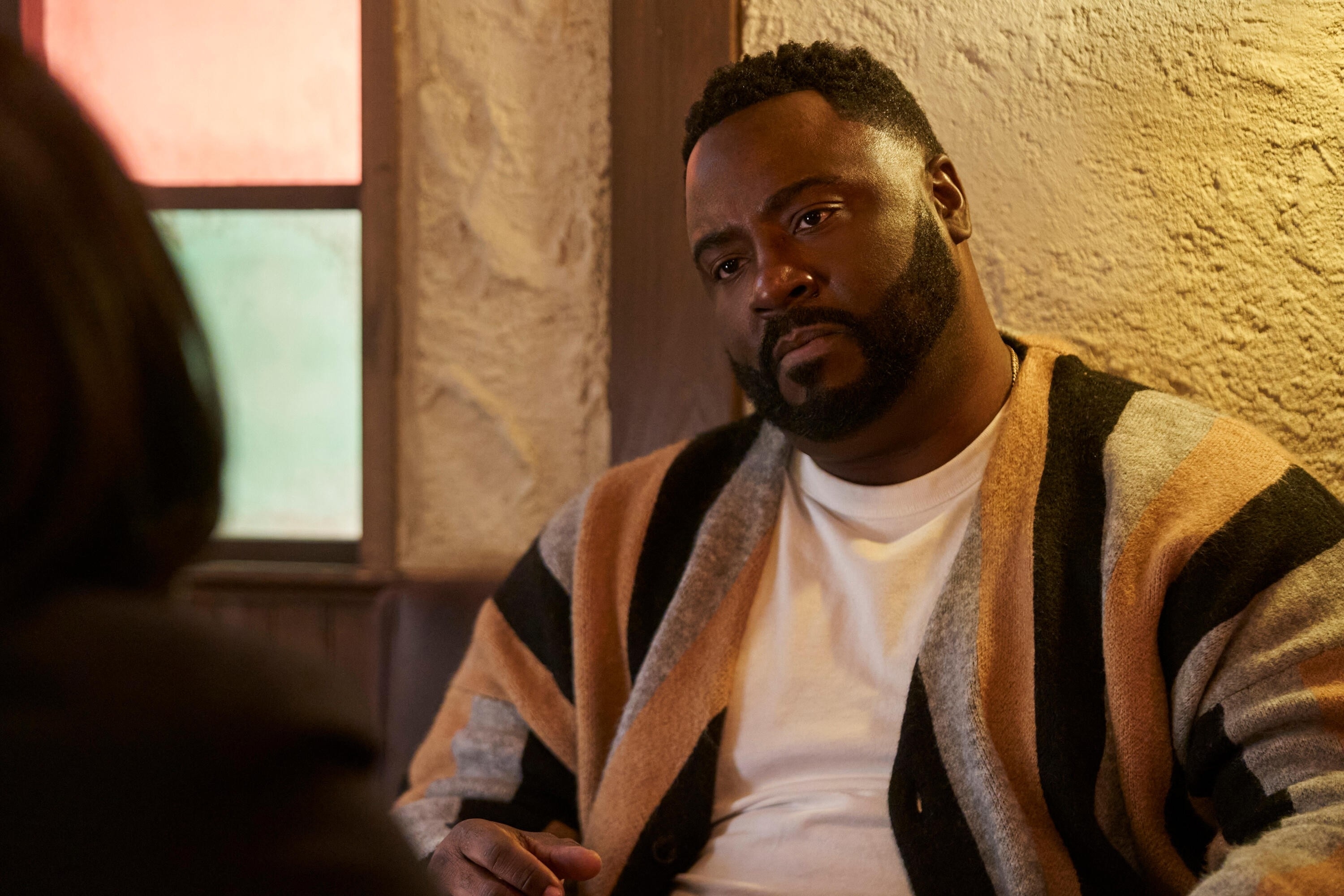 Person wearing a patterned cardigan sits indoors, facing another person, appearing engaged in conversation