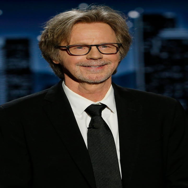 Dana Carvey in a suit with glasses, smiling at an event with blurred cityscape background