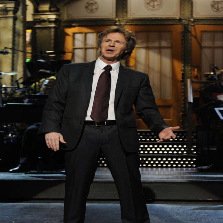 A person in a suit performing on stage with a live band in the background, likely at a comedy or entertainment show