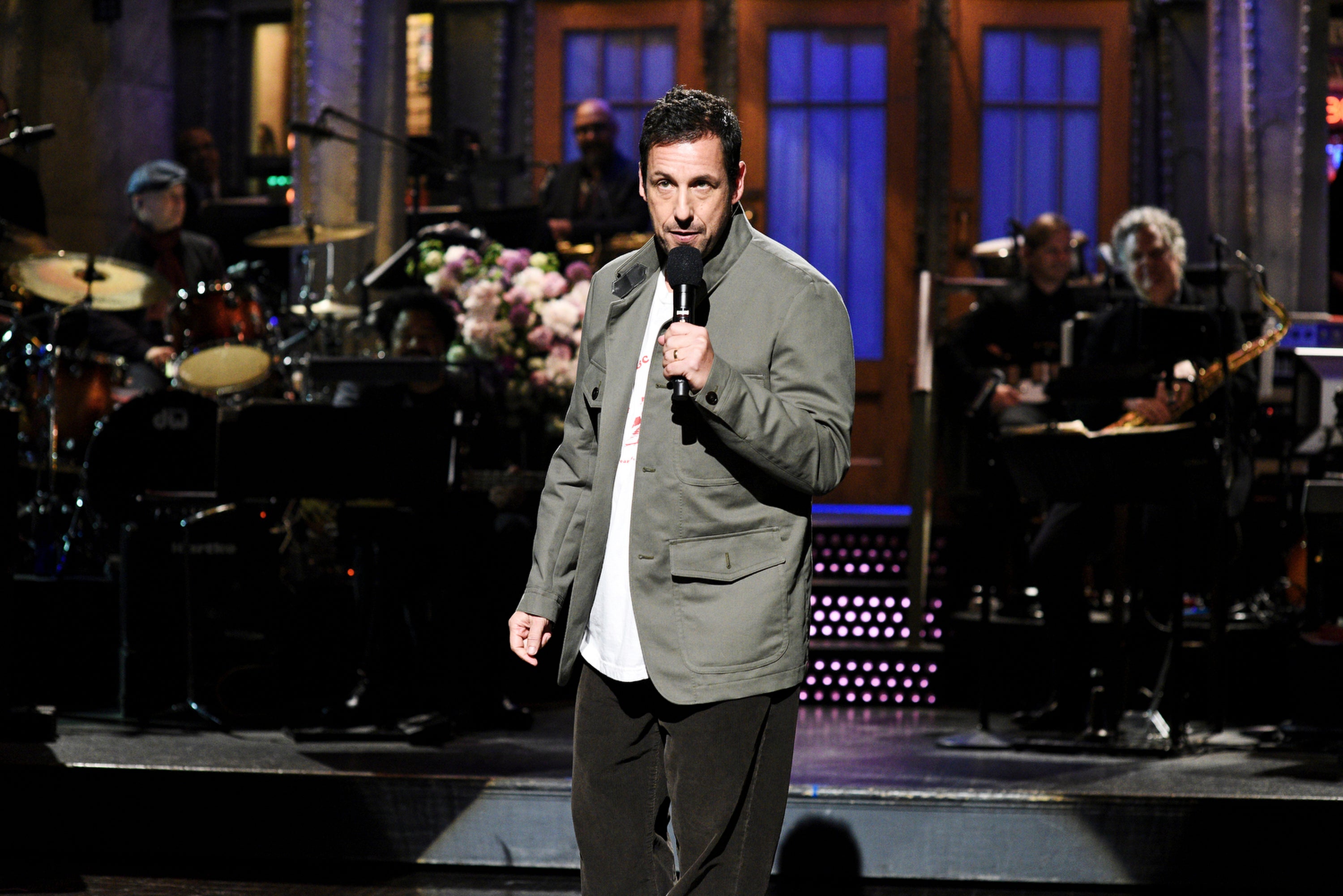 Person on stage performing with a band in the background, wearing a casual jacket and pants