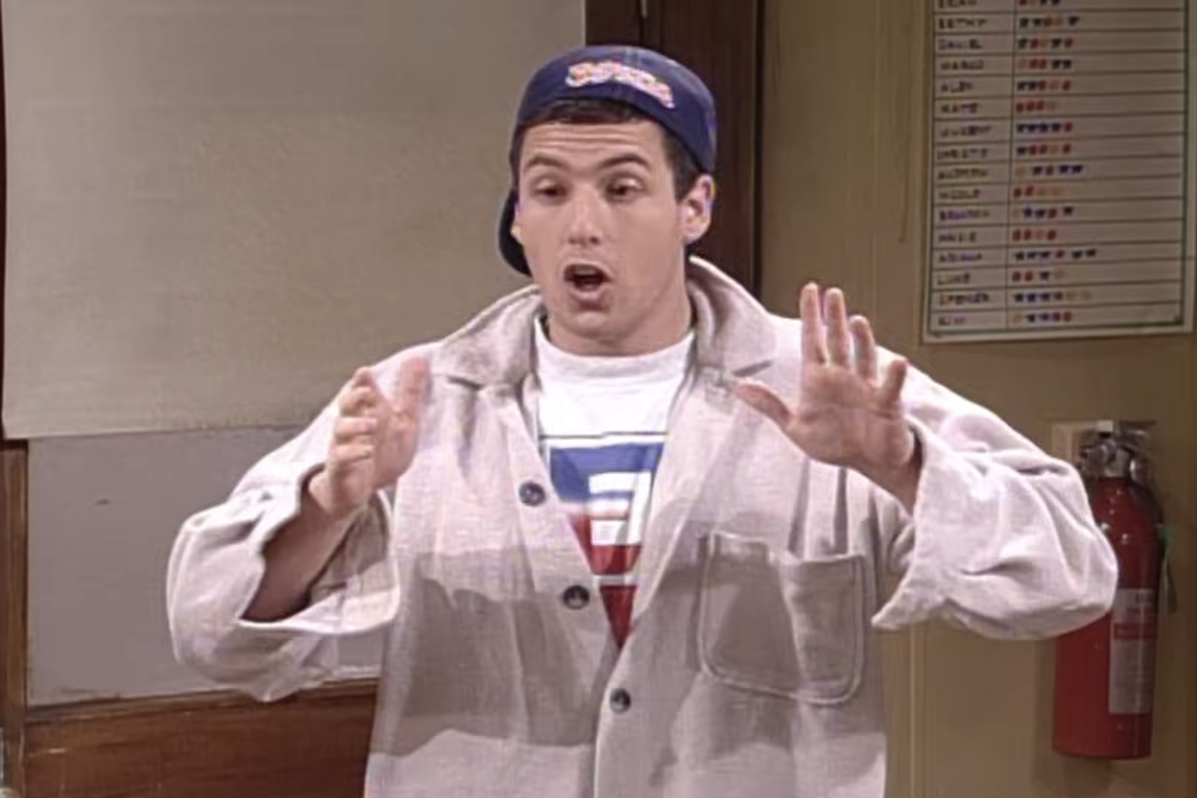 Person in casual attire and backwards cap gestures animatedly inside a room. A fire extinguisher and a chart are visible on the wall behind them