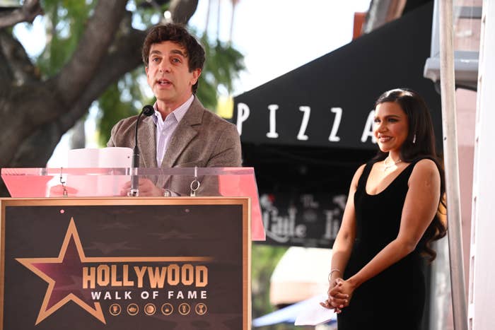A man speaks at a podium with a "Hollywood Walk of Fame" sign. A woman in a sleek black dress stands smiling beside him outdoors