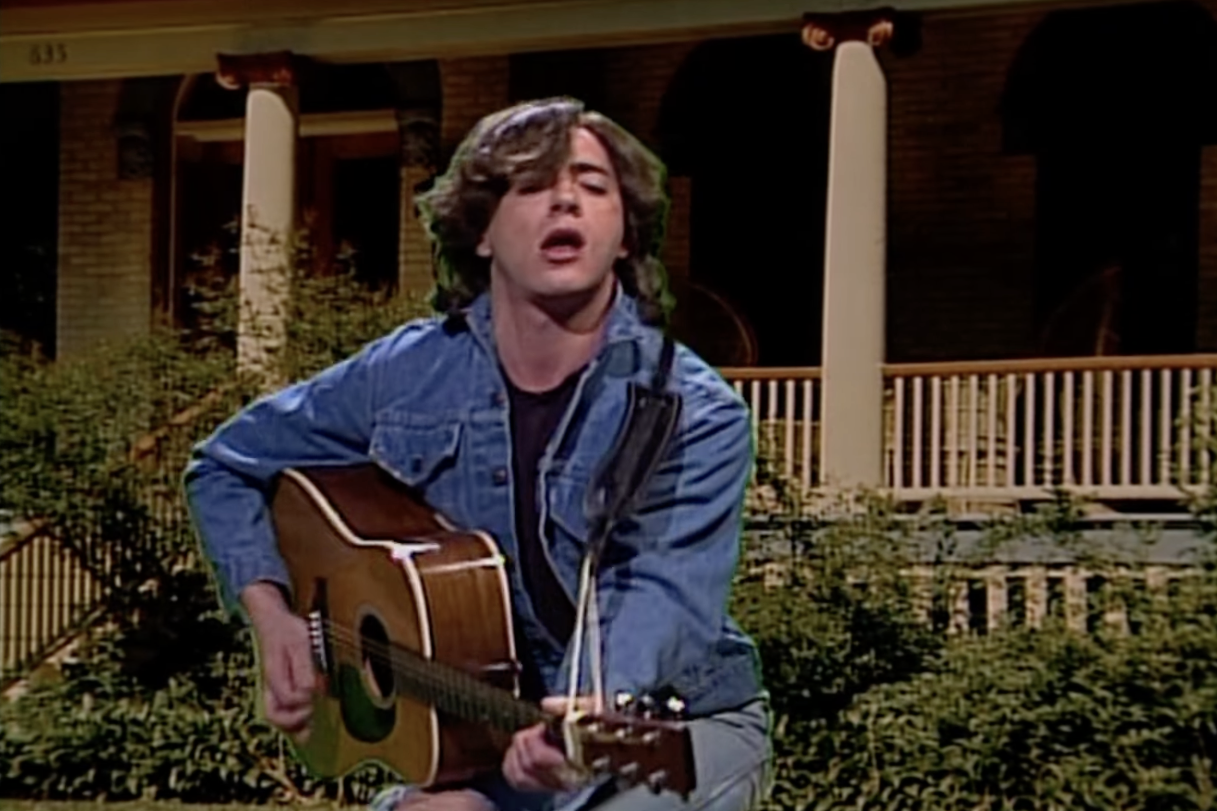 Person playing an acoustic guitar outdoors on a porch, wearing a denim jacket and shirt