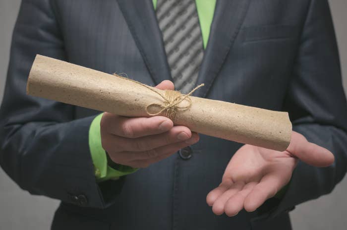 A person in a suit holds a rolled-up parchment tied with string, presenting it with one hand, possibly symbolizing an award or important document