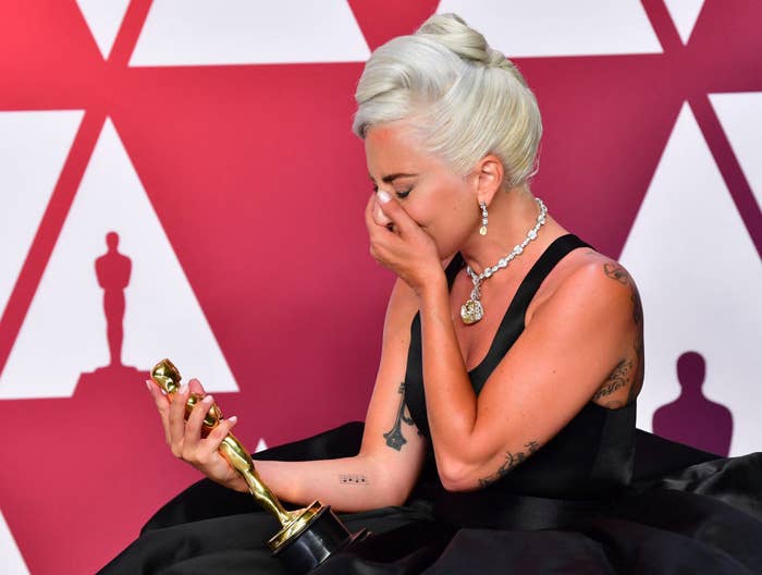 Person in elegant black gown holds an award, appearing emotional, with hand covering mouth. Triangular backdrop indicates a formal event