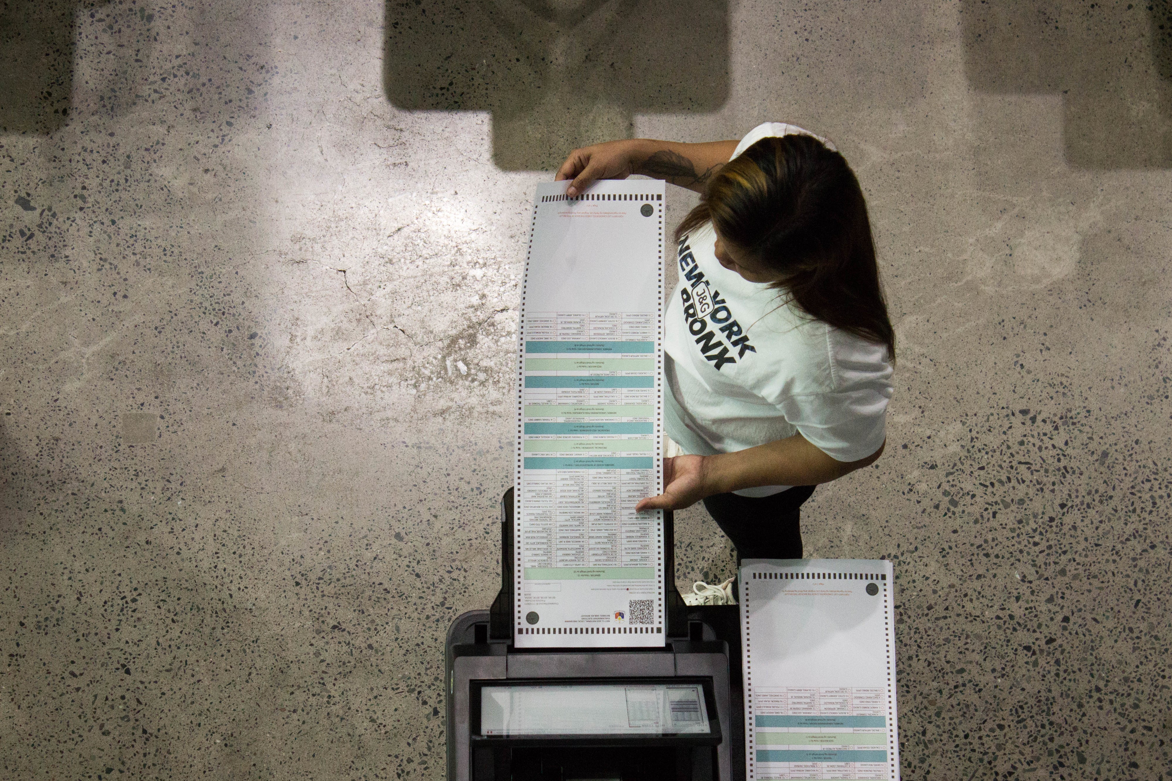 Person in "New York Economy" shirt operates a large voting machine, handling ballots. Image taken from above