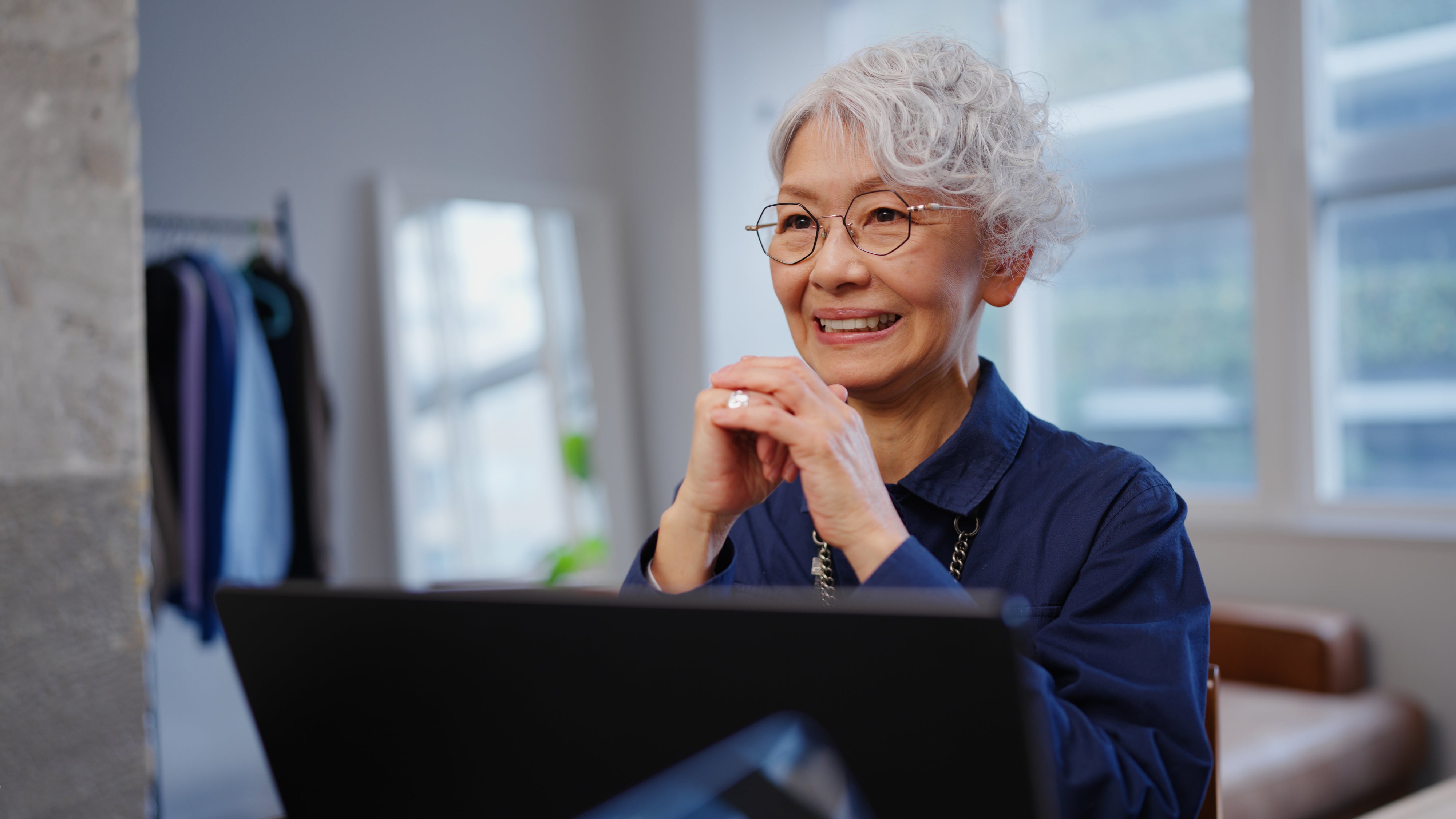 Elderly person with short hair and glasses smiling in front of a laptop, appearing engaged and thoughtful in a modern room setting