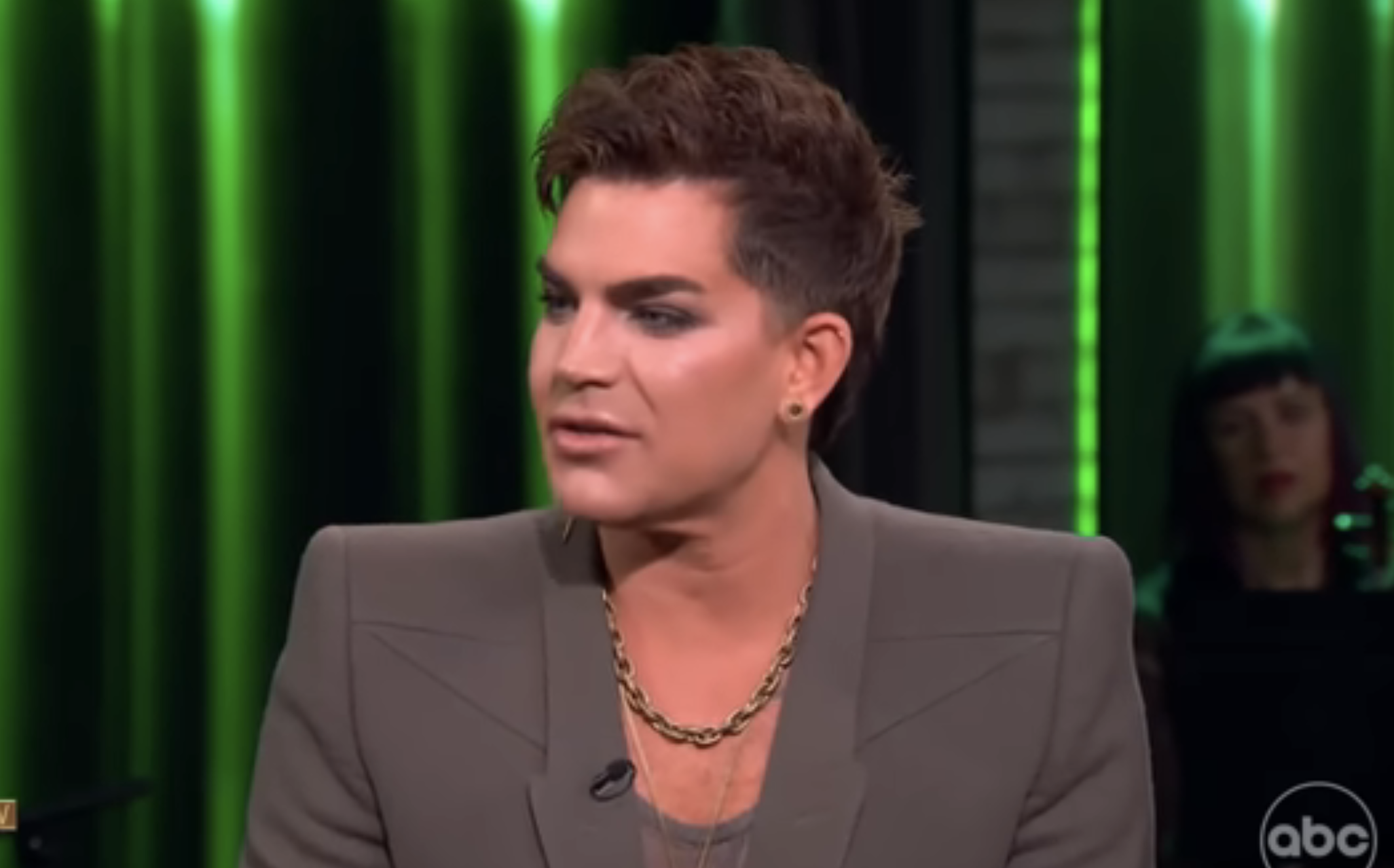 Person with styled hair, blazer, and necklace talking on a TV show set, visible audience member in the background