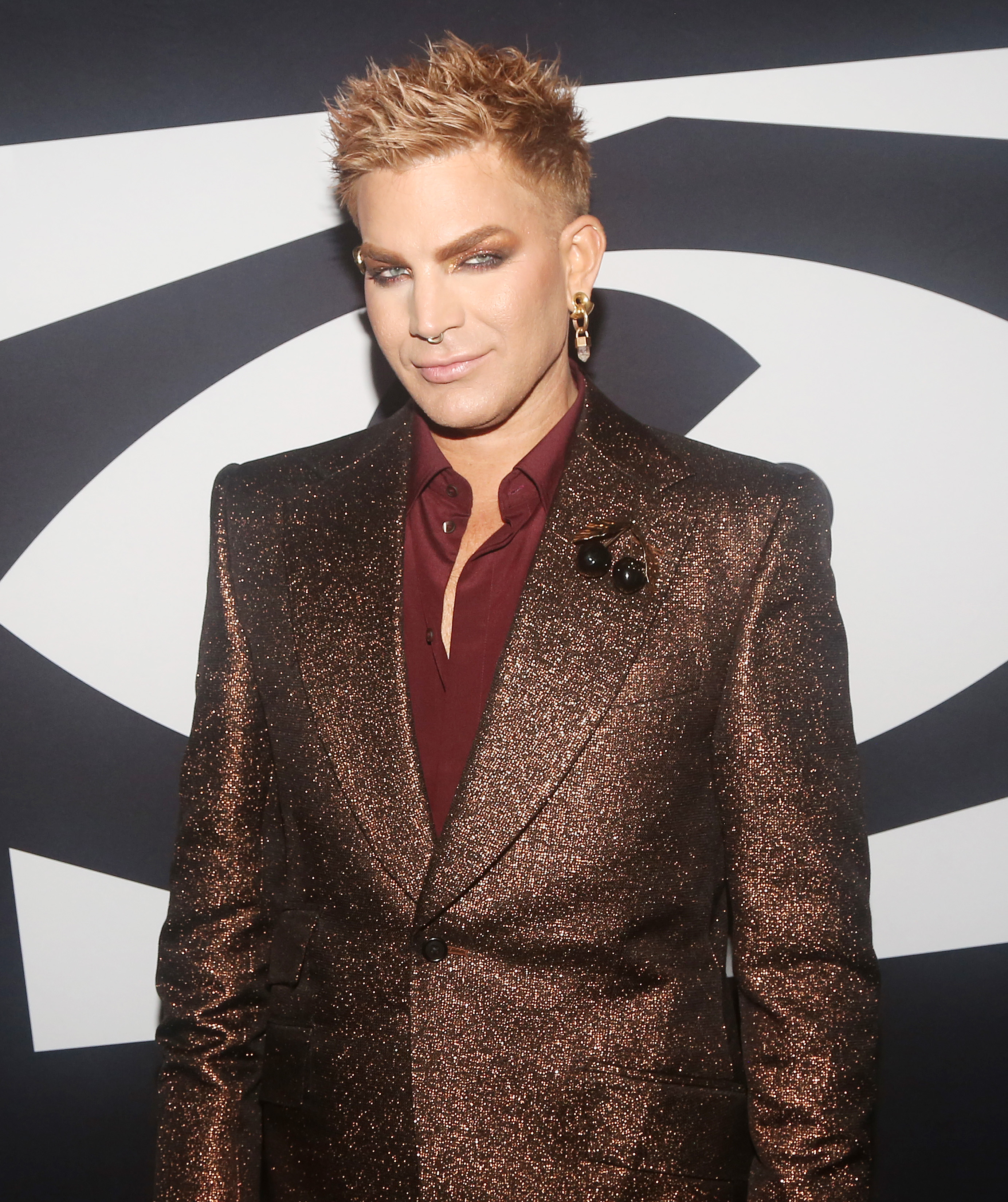 Person in a glittering suit with a deep burgundy shirt stands on a red carpet. They have styled hair and are wearing statement earrings