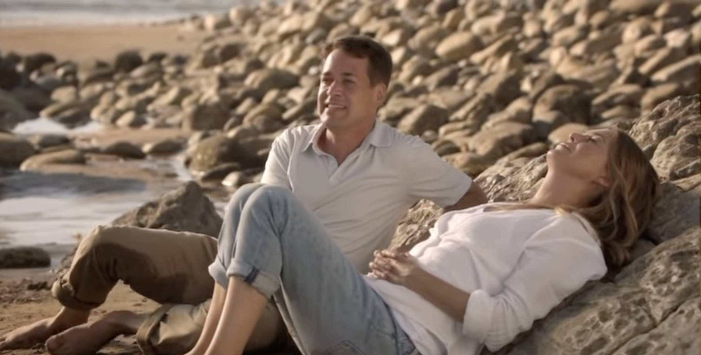 Two people sit on rocky beach, gazing contentedly towards the ocean. One leans back, relaxed, while the other sits upright