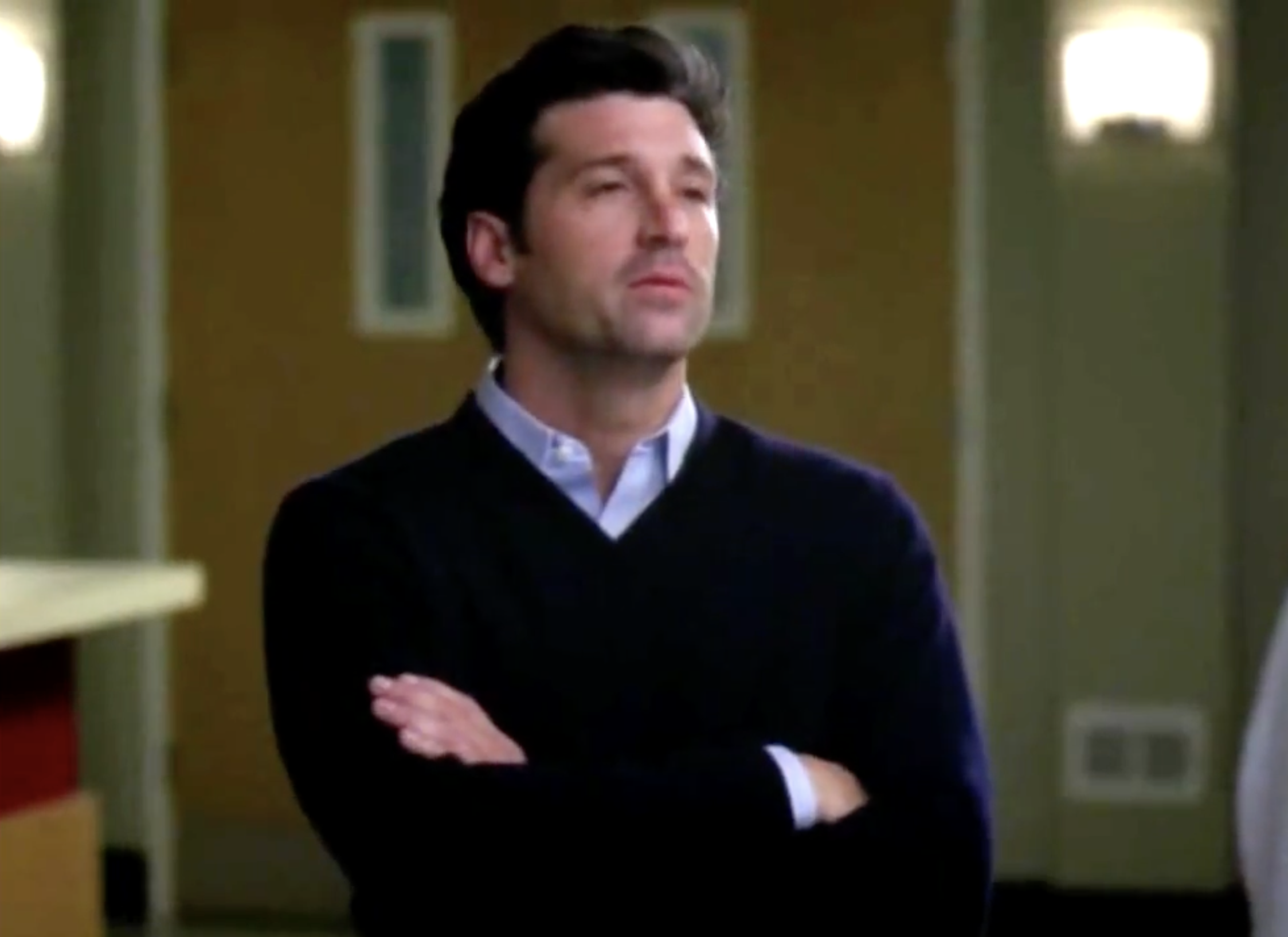 Man with crossed arms, wearing a dark sweater over a collared shirt, standing indoors with a thoughtful expression