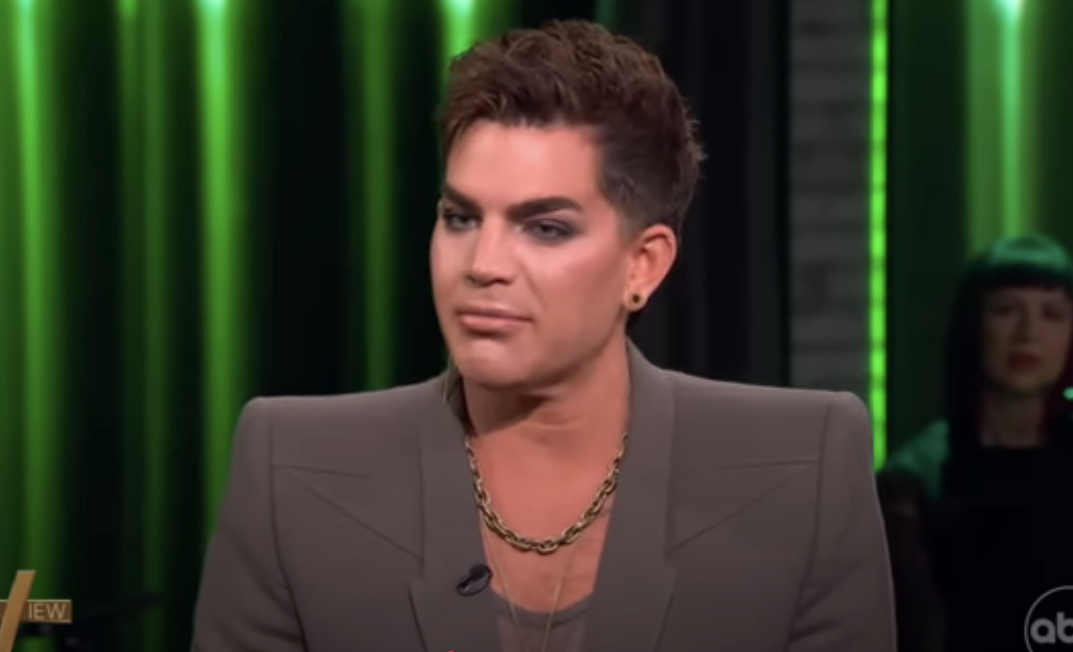 A person with styled hair and heavy makeup wears a structured blazer and chain necklace, appearing in a television interview setting