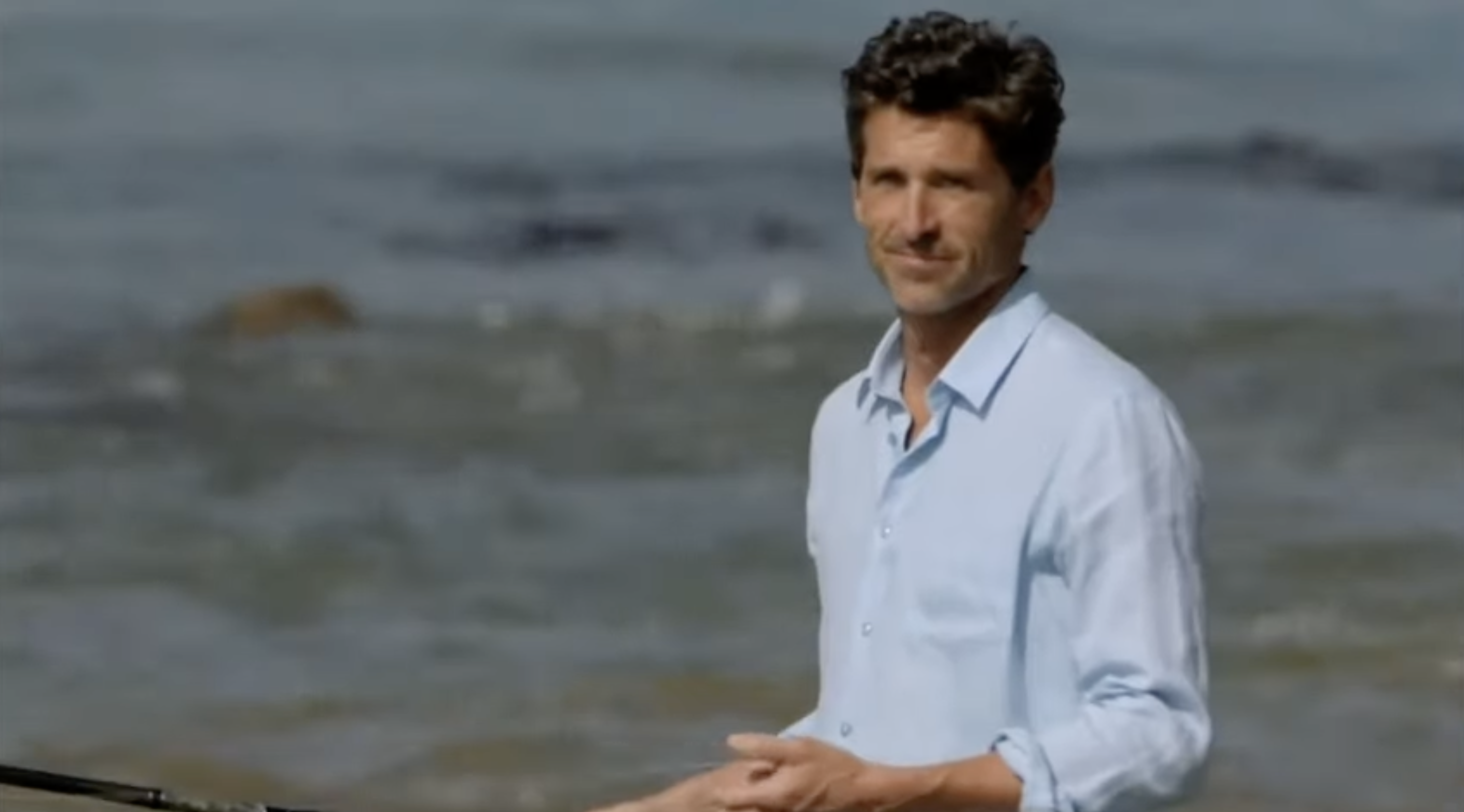 A person stands by the ocean, wearing a light, casual shirt, with waves in the background