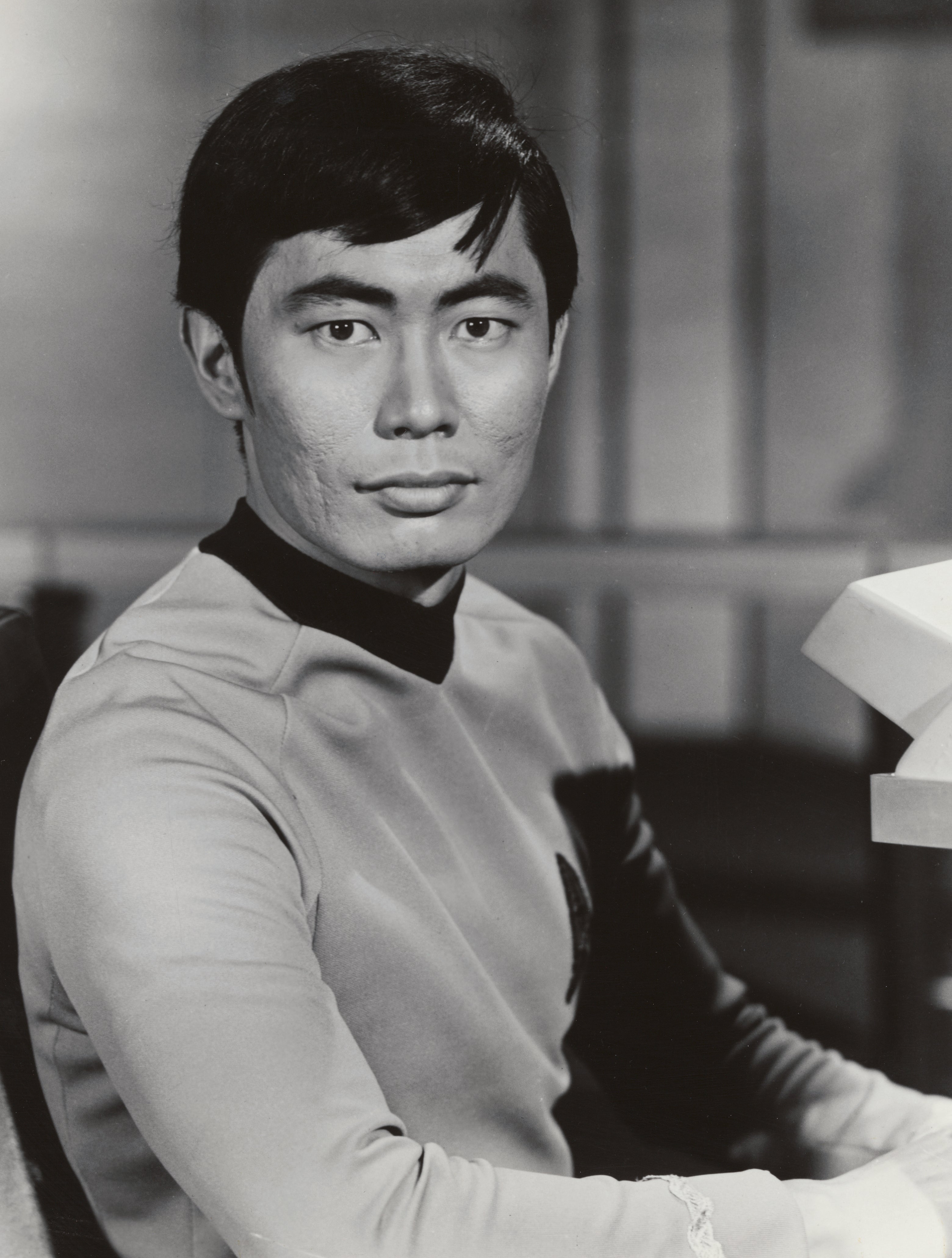 Person in a retro sci-fi uniform sits, looking towards the camera. The uniform has a high collar and an emblem on the chest